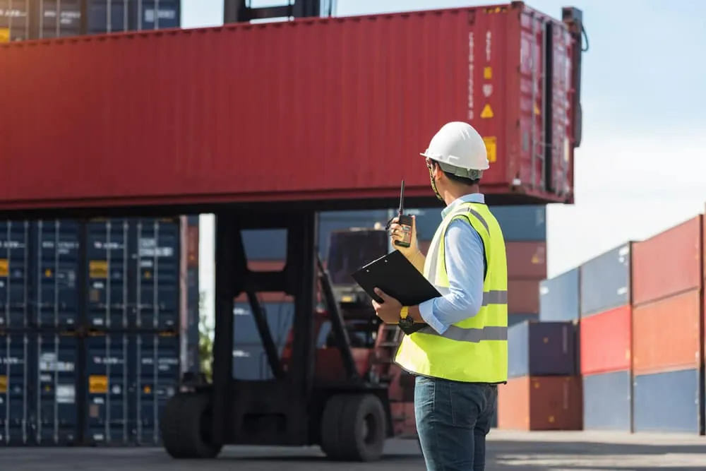 Veiligheidskundige met witte helm, geel hesje, portofoon en klembord kijkt toe terwijl een reachstacker een rode container optilt op een containerterminal